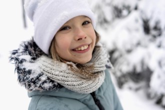 Smiling child in winter clothing with a white hat and knit scarf, surrounded by snow-covered trees.