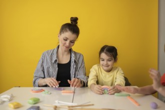 A woman and child create colorful clay shapes at a white table against a vibrant yellow background,
