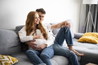 A young couple sits on a comfortable sofa, joyfully expecting their first child. The man gently