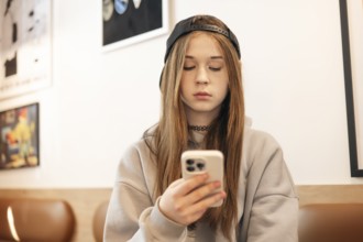 A teenage girl in a casual hoodie and cap sits indoors, focused on her smartphone. The background