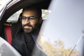 An Indian man dressed in a dark winter coat and glasses is seen smiling gently while driving his