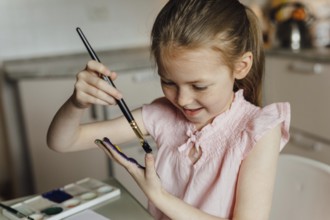 A joyful young girl in a pink top paints her hand with watercolor, showcasing creativity and fun.
