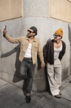 A Colombian couple taking a selfie, hand in hand. They lean against a city wall, embodying modern