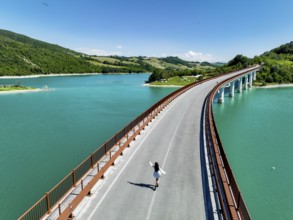 A young woman walks along a picturesque bridge overlooking a vibrant turquoise lake, surrounded by