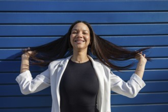 A smiling Hispanic woman in a business outfit poses confidently against a blue backdrop, reflecting