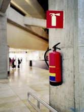 A red fire extinguisher mounted on a concrete wall in an airport terminal. A safety sign above it