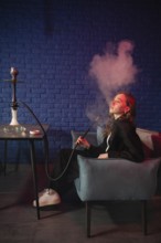 A woman enjoys a hookah in a modern lounge against a blue brick wall. Smoky clouds create a serene