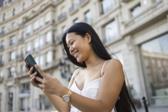 A young Chinese woman smiling while using her smartphone on a sunny summer day in the city. The