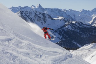 A snowboarder wearing bright red gear carves down a snowy mountain slope. The backdrop features