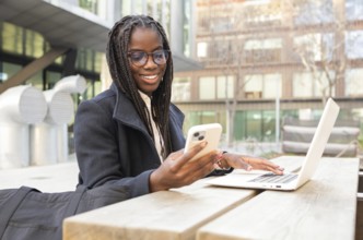A young African American businesswoman with braids is smiling as she uses her smartphone and laptop