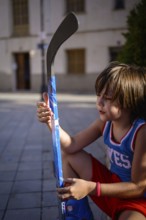 A young child sits on a paved street, holding a vibrant blue hockey stick. The scene captures a