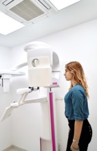 A young woman stands in a dental clinic, preparing for a panoramic dental X-ray. The advanced