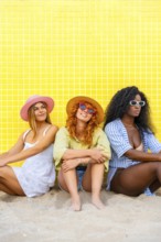 Three young women wearing summer clothes and sunglasses are sitting on the sand against a yellow
