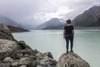 A woman stands on a rocky outcrop, taking in the serene autumn beauty of Mount Cook in New Zealand.