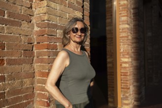 A joyful mature woman in casual attire stands against a brick wall, basking in the warm sunlight.
