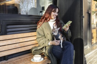 A woman enjoys a coffee while sitting on a bench outside a cafe, holding her phone and smiling at