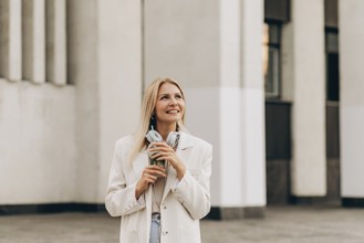 A smiling woman stands outside in an urban setting, holding a drink and headphones. She wears a