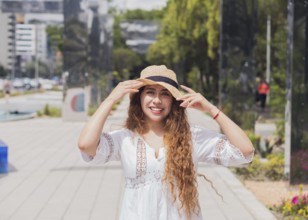 Young American Latin woman in a casual white dress and straw hat, smiling brightly in an urban park