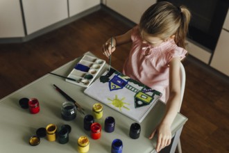 A child, dressed in a pink top, is intently painting a vibrant scene of houses and a sun using