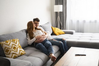 A young couple sits lovingly embraced on a gray sofa, hands gently resting on the woman's pregnant