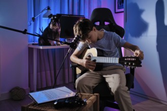 A young boy plays an acoustic guitar in a modern room with colorful lighting. A dog sits nearby on