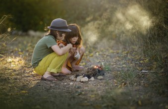 Two children sitting closely outdoors by a small campfire, surrounded by nature. They are focused