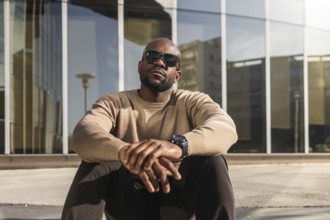 A confident cuban man wearing sunglasses and a casual beige sweater sits outdoors against a modern