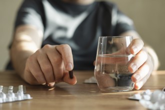 Man sits at a table, holding a pill in one hand and a glass of water in the other. The scene
