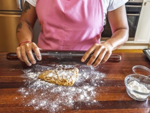 A woman in a pink apron uses a dark rolling pin to flatten dough on a floured wooden counter. A