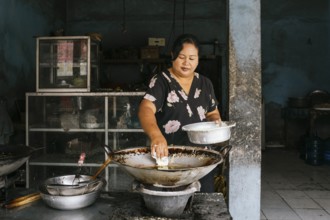 A woman prepares traditional food in a rustic kitchen of Bali, focusing intently as she cooks over