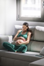 A pregnant woman peacefully sitting on a sofa in a sunlit room, dressed in a comfortable green yoga