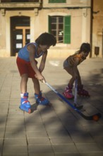 Two children play street hockey on a sunny day, wearing roller skates in an urban setting. The