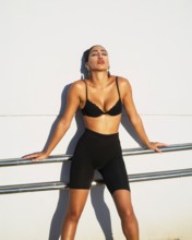 A confident woman in a fashionable black outfit poses against a white wall illuminated by sunlight.