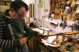 A luthier meticulously crafts a bass guitar neck in a workshop filled with tools. The focus is on