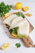 Fresh squid prepared for cooking, surrounded by herbs and lemon, placed on a wooden chopping board