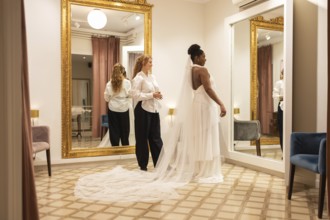 A bride admires her reflection in an elegant boutique fitting room. An assistant helps with her