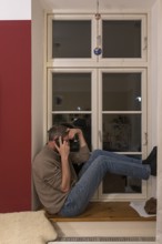 Young man sitting on the windowsill while talking on the phone, Mecklenburg-Vorpommern, Germany