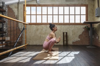 A woman wearing pink active wear is practicing yoga indoors on a sunny day. She is in a squatting
