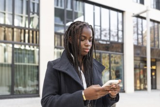 A young African American woman with braids, dressed in a business coat, is using a smartphone while