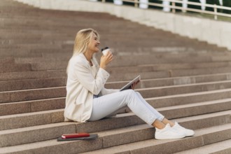 A businesswoman sits on outdoor steps with a tablet, enjoying a coffee. Dressed casually in white