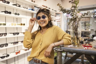 A cheerful Latin woman wearing stylish sunglasses poses in a trendy eyewear shop, showcasing a