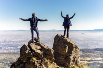 Two friends stand triumphantly on a mountain peak, facing a clear blue sky and expansive landscape,