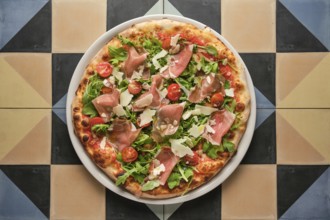 Overhead view of a whole pizza topped with prosciutto, arugula, and tomatoes, served on a vibrant,