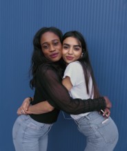 Two young women embrace in a joyful, friendly hug against a vibrant blue background. Their radiant