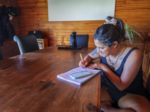 A woman is writing in a notebook at a large wooden table, surrounded by warm decor in Australia.