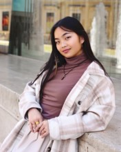 A young chinese woman with long dark hair wearing a plaid coat and brown turtleneck leans casually