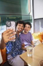 A joyful, multiethnic LGBTQ+ couple sits in a cozy cafe, smiling as they take a selfie. Sunlight