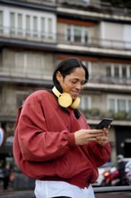 A man with long hair and large glasses wearing a red jacket and yellow headphones is outside