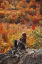 A woman sits on a rocky outcrop wrapped in a blanket, gazing at the stunning autumn foliage of