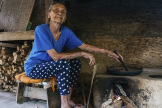 An elderly woman in a rustic kitchen prepares balinese coffee on a traditional wood fired stove.
